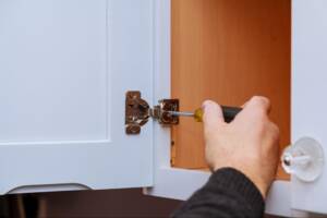 A close-up of a man's hand adjusting a cabinet door hinge on kitchen cabinets.