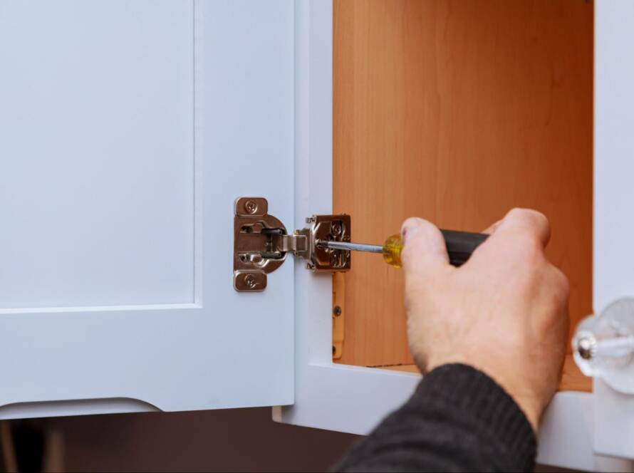 A close-up of a man's hand adjusting a cabinet door hinge on kitchen cabinets.
