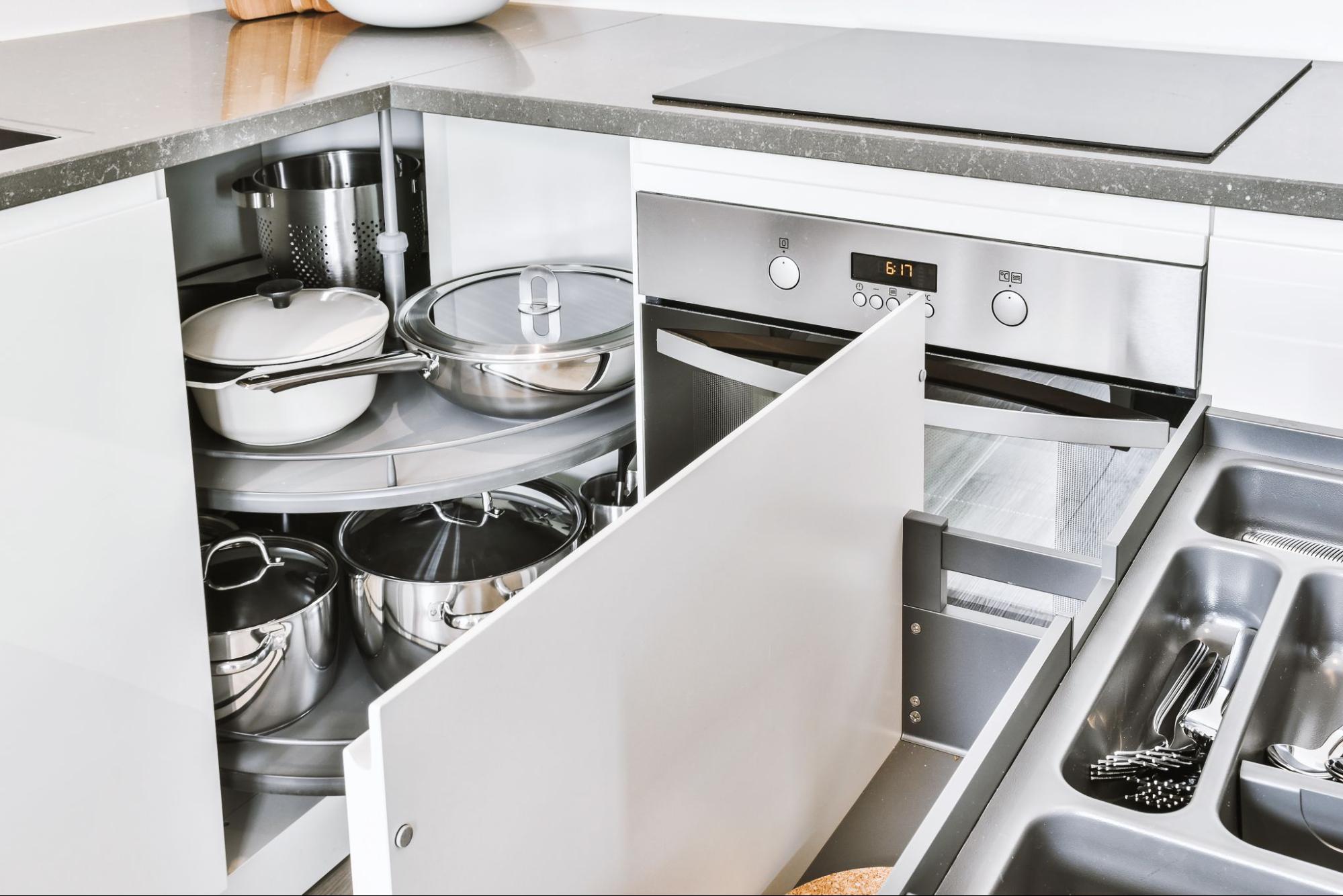 A close-up of a kitchen corner cabinet with storage drawers, neatly organized pots, and pans seamlessly integrated into white cabinets alongside an oven.