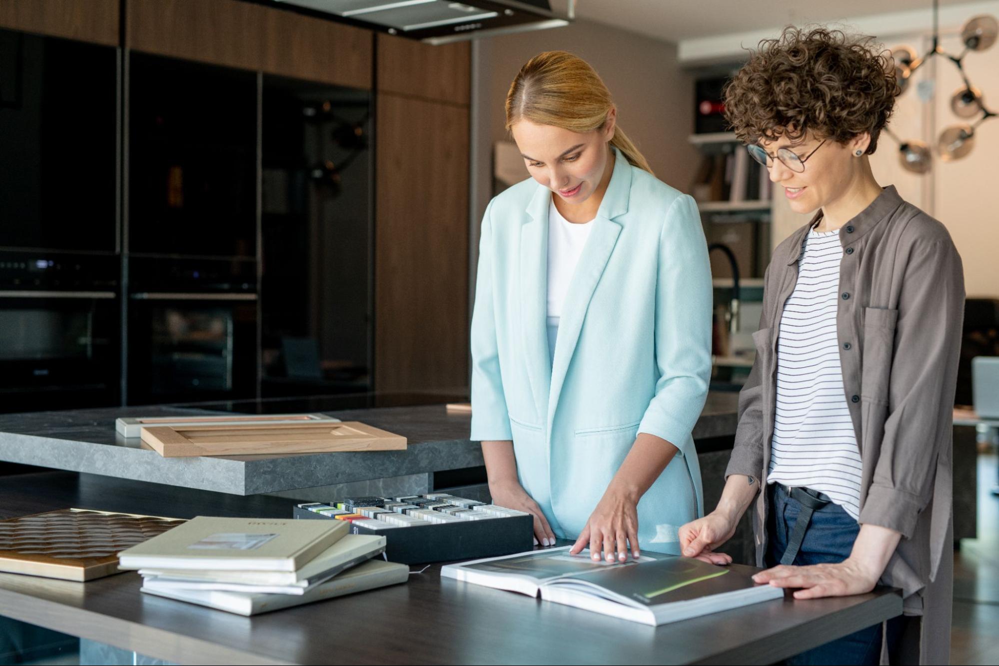 A store assistant wearing a teal blazer over a white top guides the client, dressed in a striped white and black t-shirt under a gray button-up shirt on different cabinet styles available in the store. 