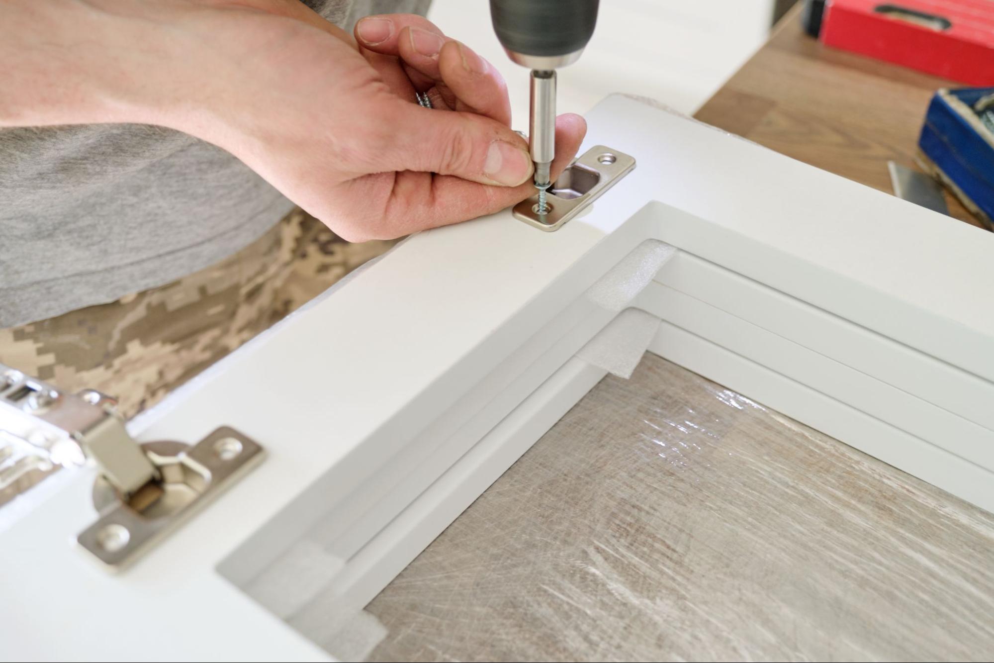 A close-up of a worker's hand adjusting cabinet hinges with professional tools on the floor.