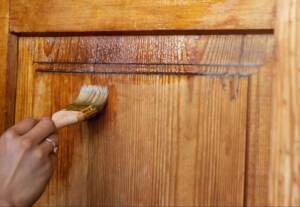 Close-up of a girl's hand staining a wooden kitchen cabinet door with a brush.