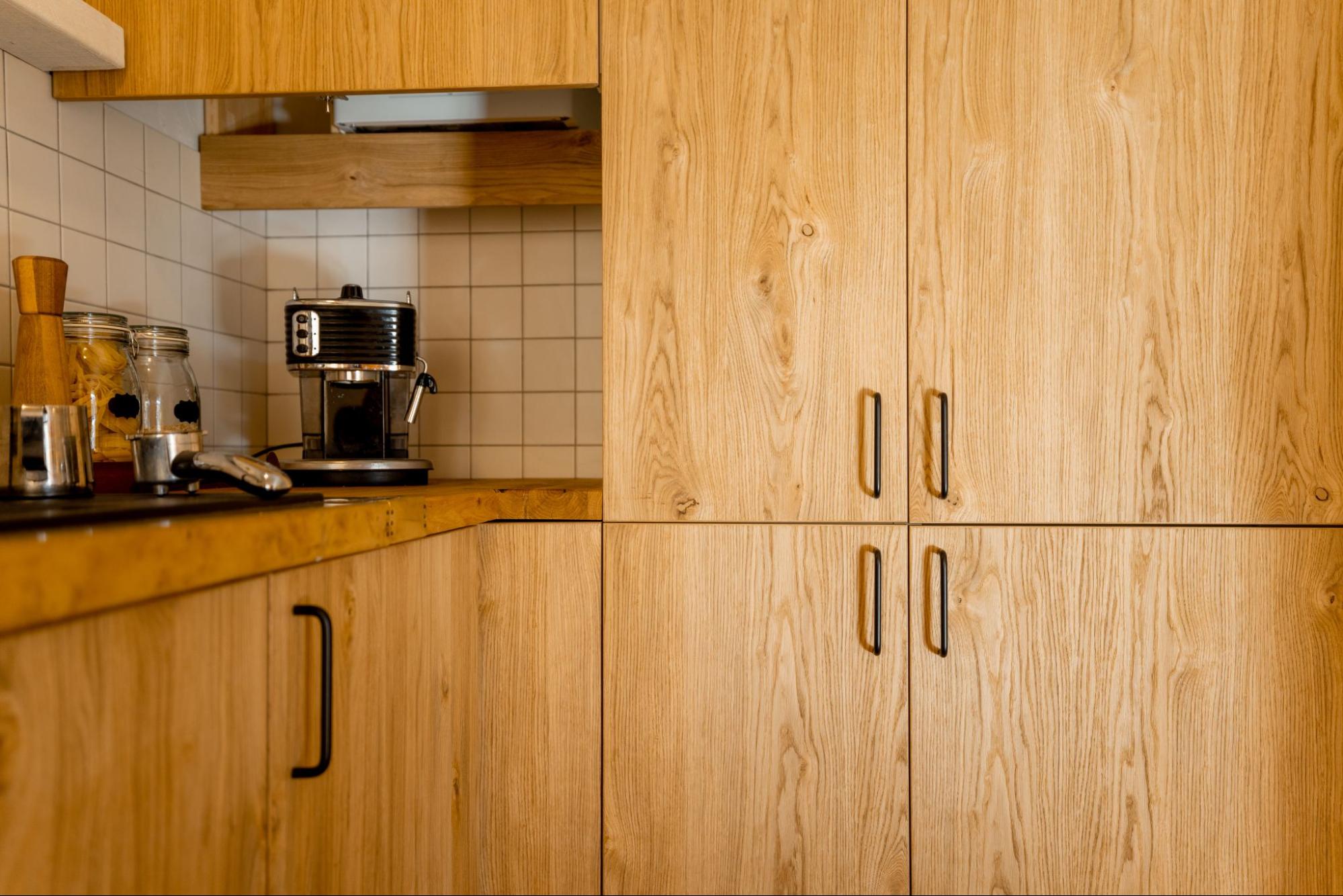Kitchen wooden cabinets with a coffeemaker and neatly arranged coffee ingredients in jars underneath, positioned in the corner against white tiles.